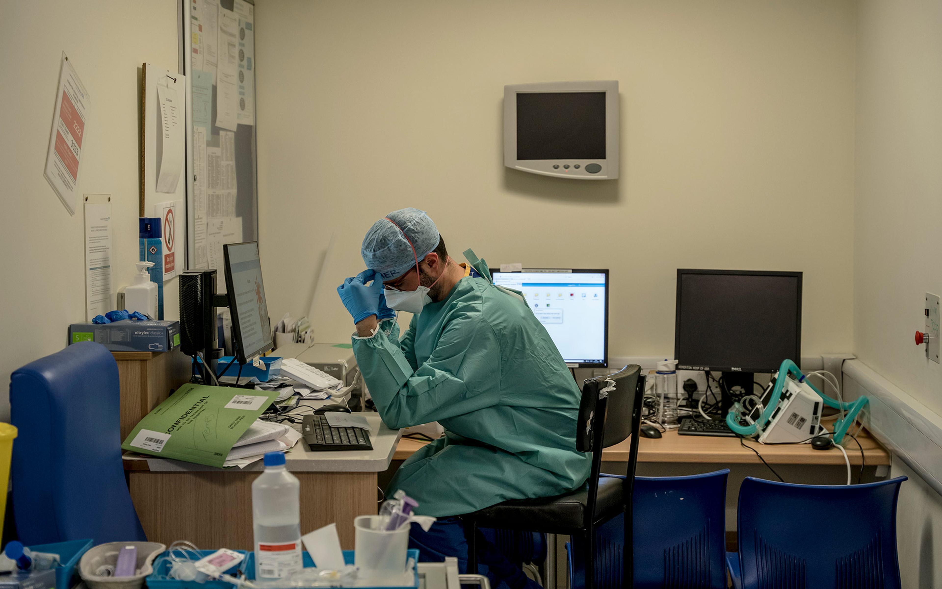 A weary looking medical staff member in scrubs and face mask sits at a desk in a hospital room surrounded by medical paraphernalia